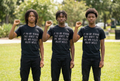 Three boys with serious, unsmiling expressions and raised fists on a grassy lawn